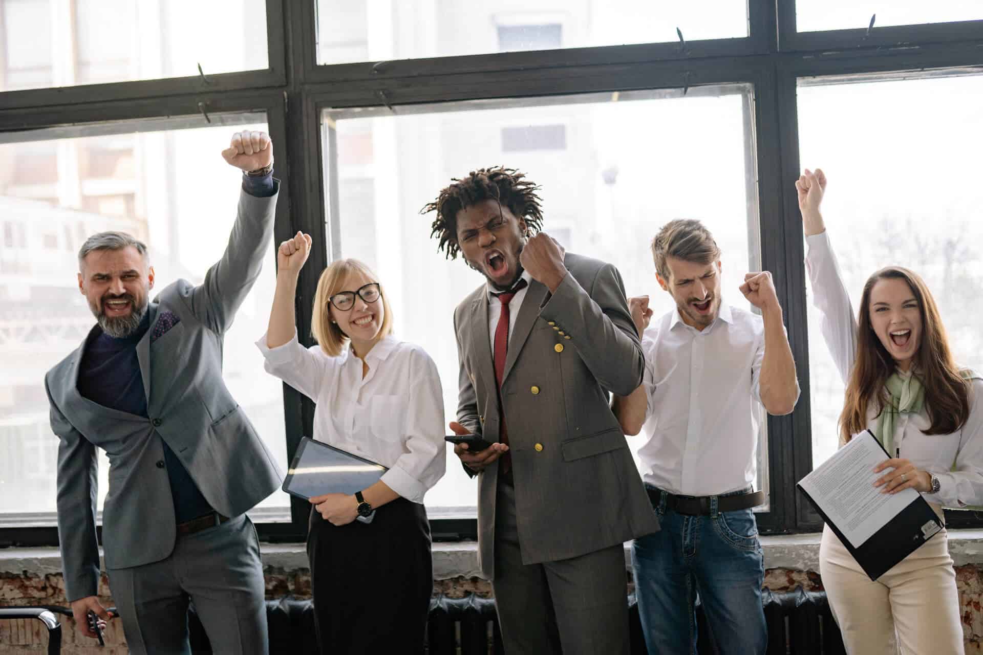 Diverse group of five people in business attire celebrating enthusiastically in a bright office setting.