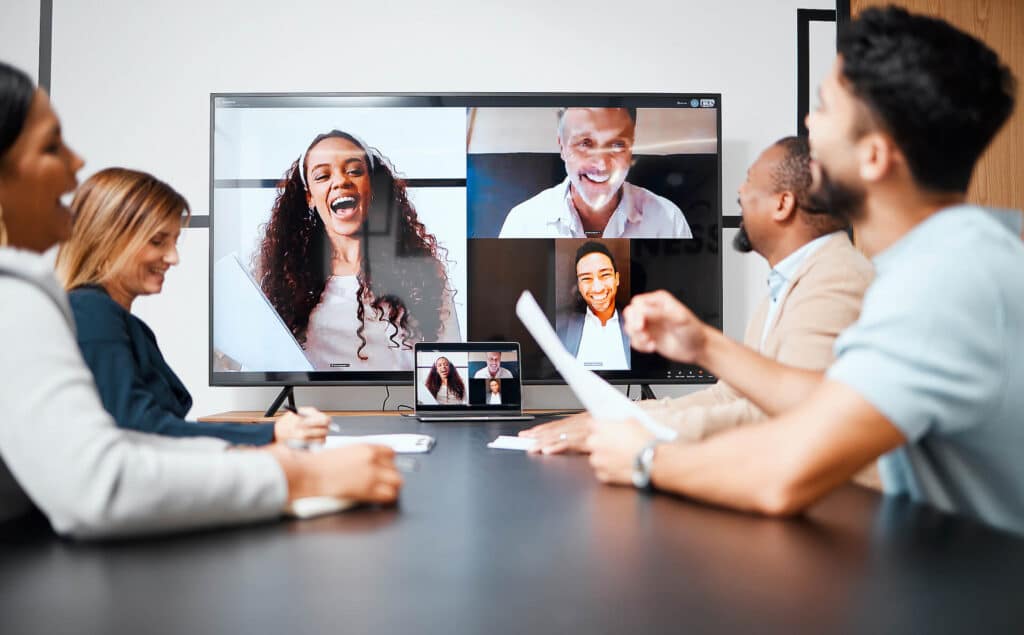People in a meeting room smiling at a screen, having a video conference with colleagues displayed on the monitor.