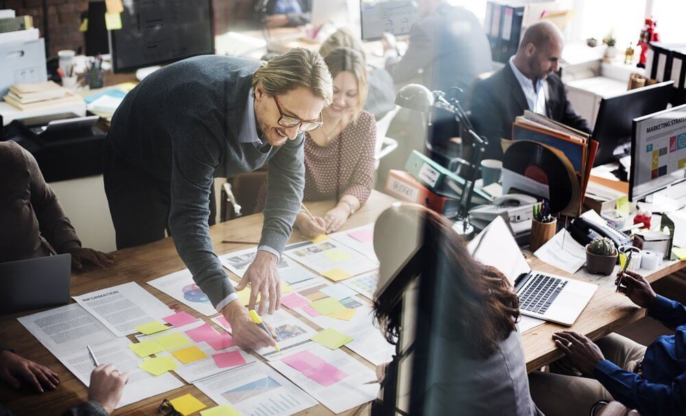 Team collaborating around a table with papers and sticky notes in a modern office setting.