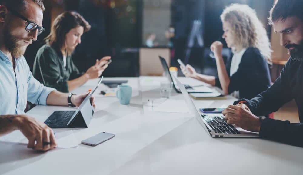 People working on laptops and phones at a shared table in a modern office with a blurred background.