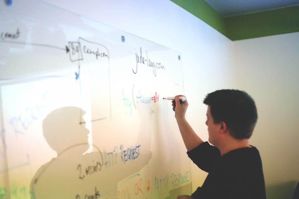 Person writing on a glass board with notes and diagrams in a bright room.