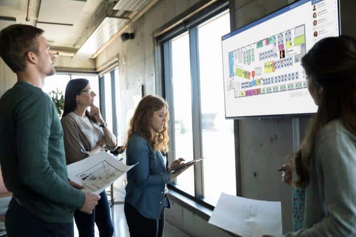 Four people discussing a floor plan displayed on a wall-mounted screen in a modern office setting.