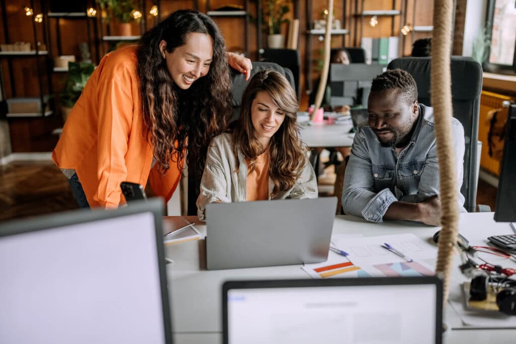 Three people happily collaborate around a laptop in a modern office setting.