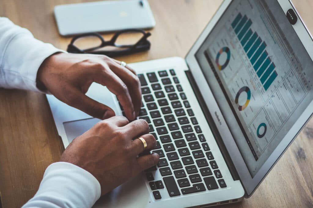 Hands typing on a laptop displaying graphs and charts, with glasses and a smartphone on the table.