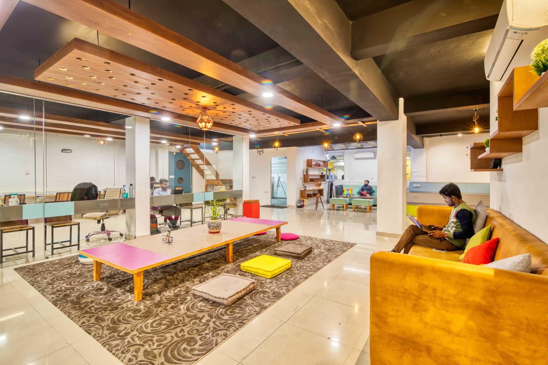 Modern open office space with a lounge area, low tables, cushions, and desks, featuring warm lighting.