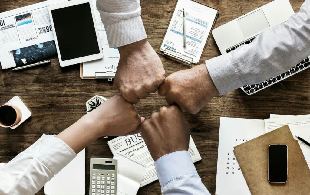 Four hands fist-bumping over a desk with papers, a tablet, keyboard, and other office items.