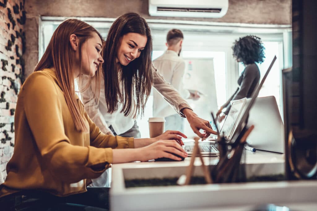 Two women smiling and working on a laptop at a desk, with two colleagues discussing in the background.