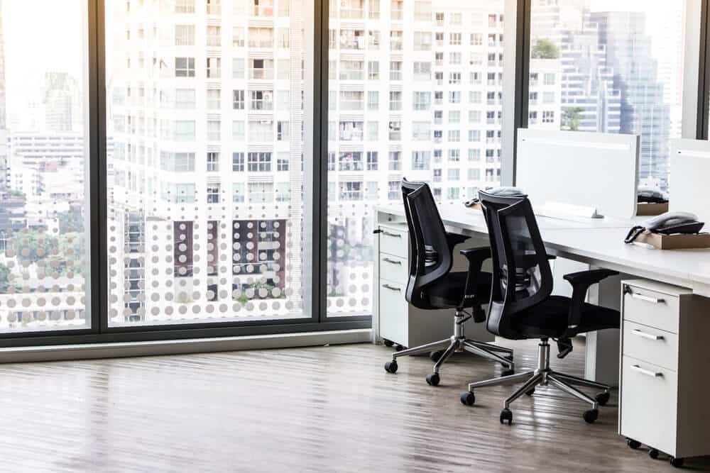 Modern office with desks and chairs by large windows overlooking a cityscape with tall buildings in daylight.