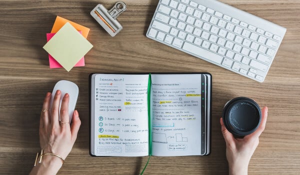 Hands holding a mouse and a coffee cup, with an open notebook, keyboard, and colorful sticky notes on a desk.