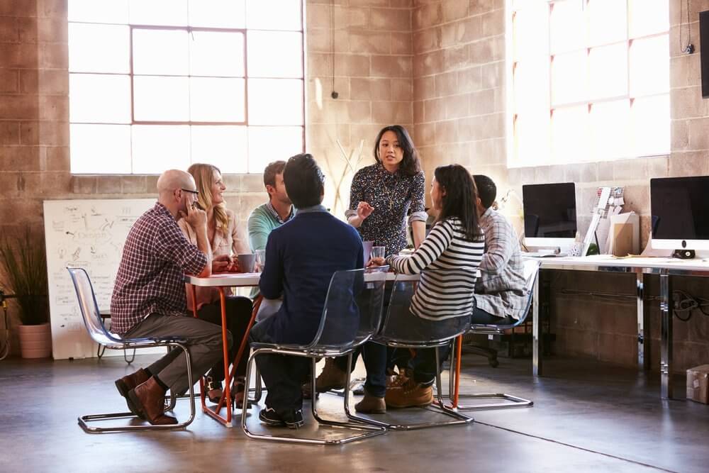 Group of people having a meeting around a table in a modern office with large windows and computer stations.