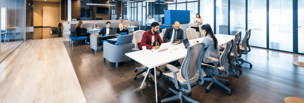 Open office space with people working at desks and in lounge areas, featuring glass walls and modern furnishings.