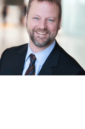 Smiling man in a suit with a light blue shirt and patterned tie, standing in a bright indoor setting.