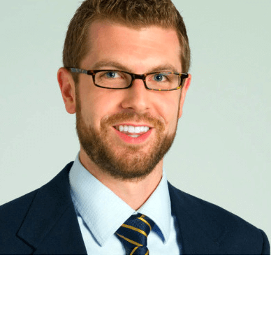 Man with glasses, beard, and a blue suit with a striped tie, smiling against a plain background.