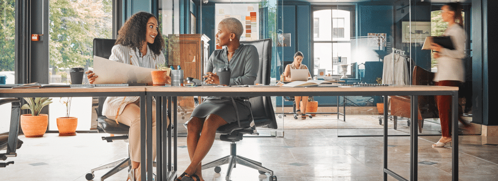 Two women seated at a desk in an office, talking and smiling. Other people work in the background.