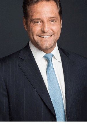 Smiling man in a dark suit with a white shirt and light blue tie against a dark gray background.