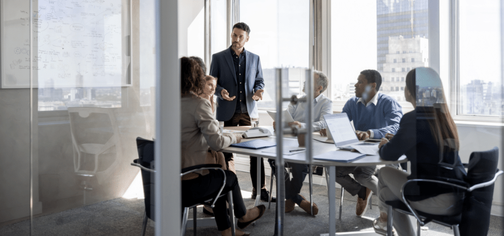 A group of people in a modern office meeting room with city views, one person stands speaking to seated colleagues.
