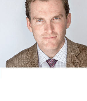Man in brown checkered suit with a subtle smile against a light background.