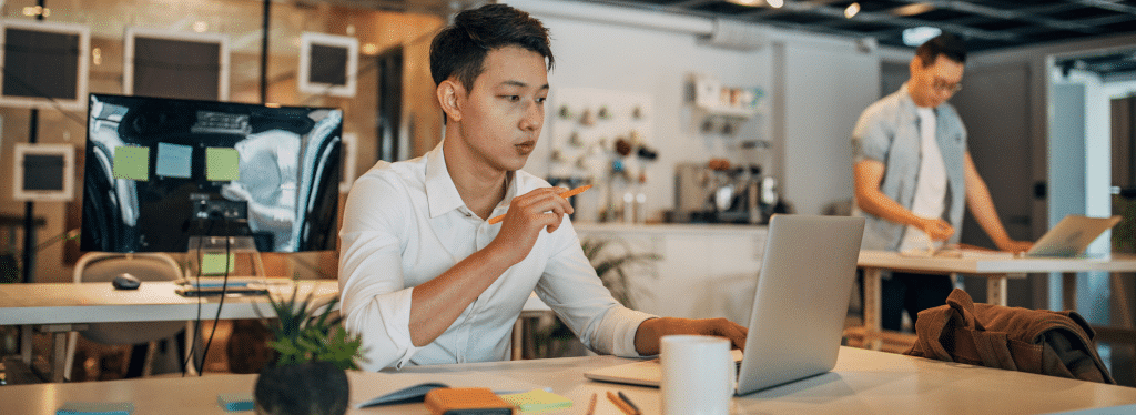 Two people working in a modern office; one at a laptop and the other standing with a notebook.